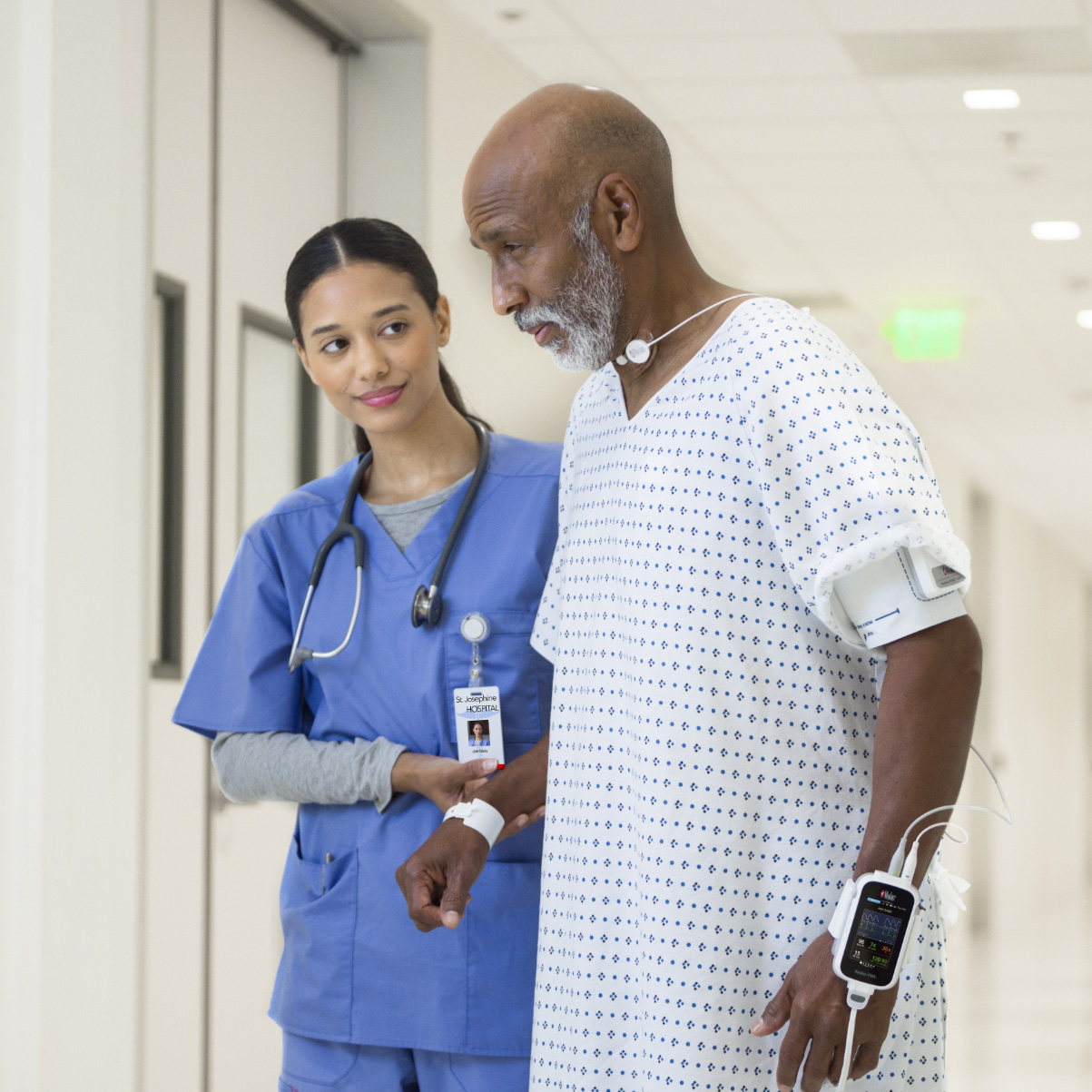 Dark-skinned patient being helped by nurse in scrubs walking down the hall in hospital gown wearing a Masimo Radius VSM monitor. 