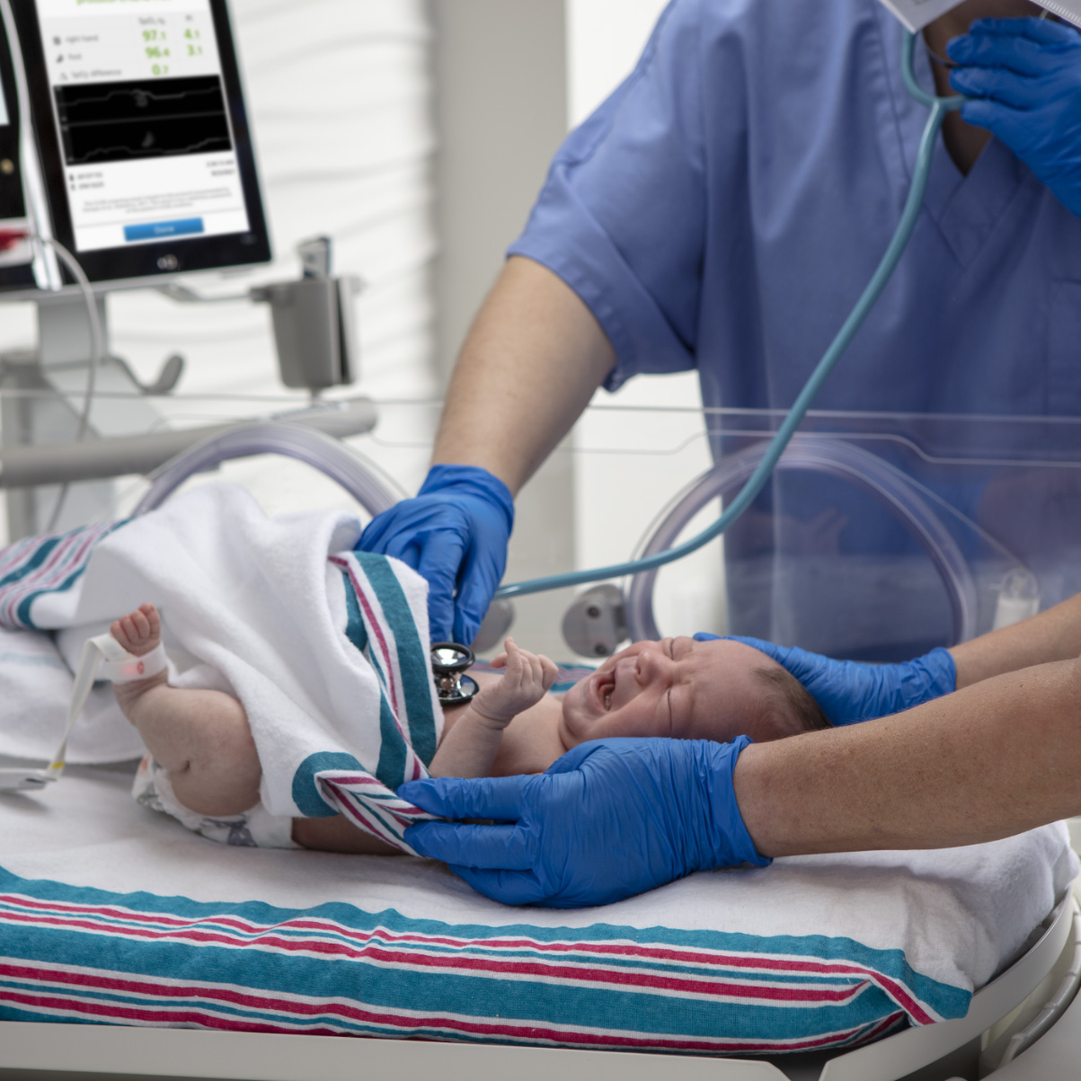A newborn infant being screened by clinicians using a stethoscope and monitored with a Masimo Newborn pulse oximetry sensor.